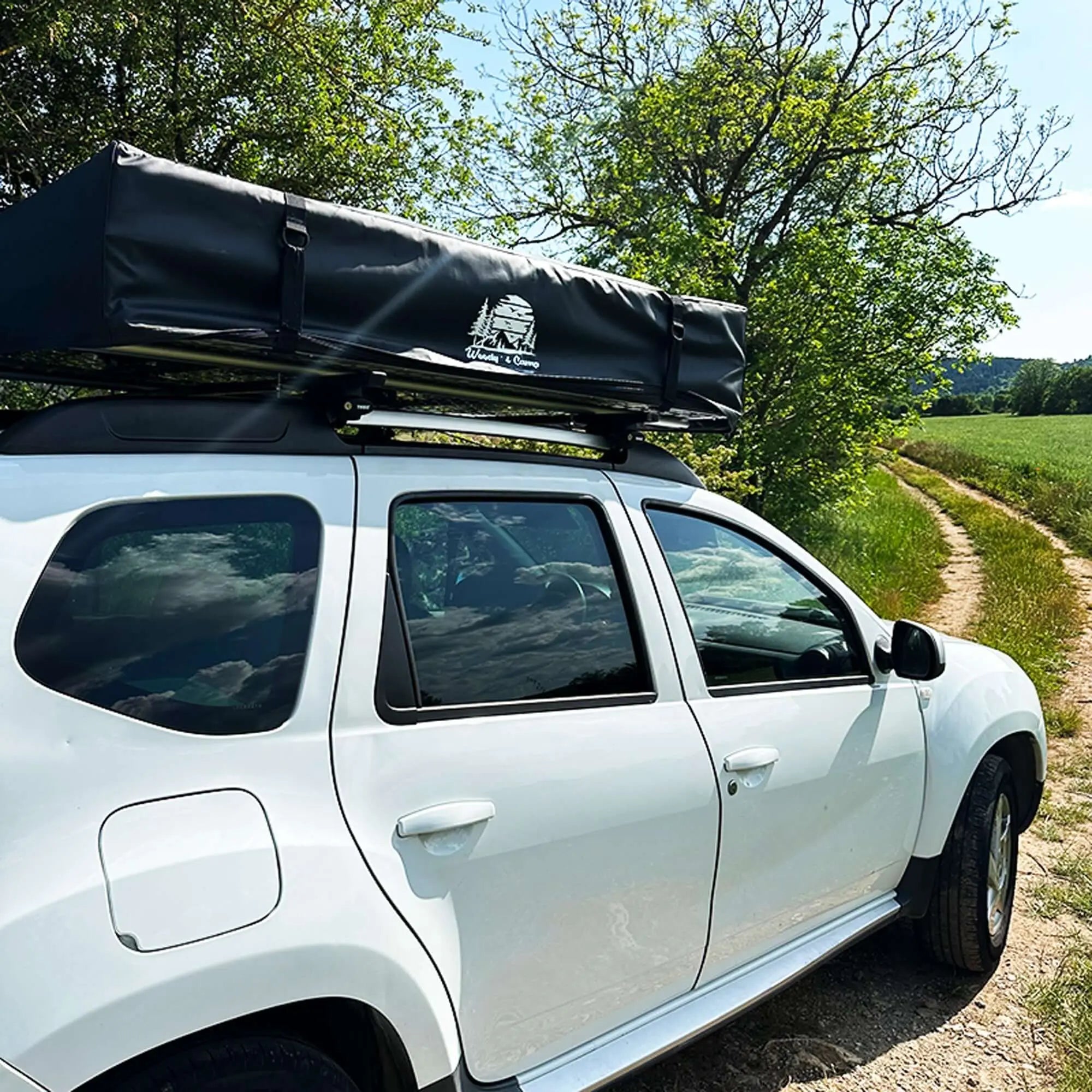 Car with soft rooftop tent for camping, ideal for 4-5 people, parked on a rural path surrounded by lush greenery.
