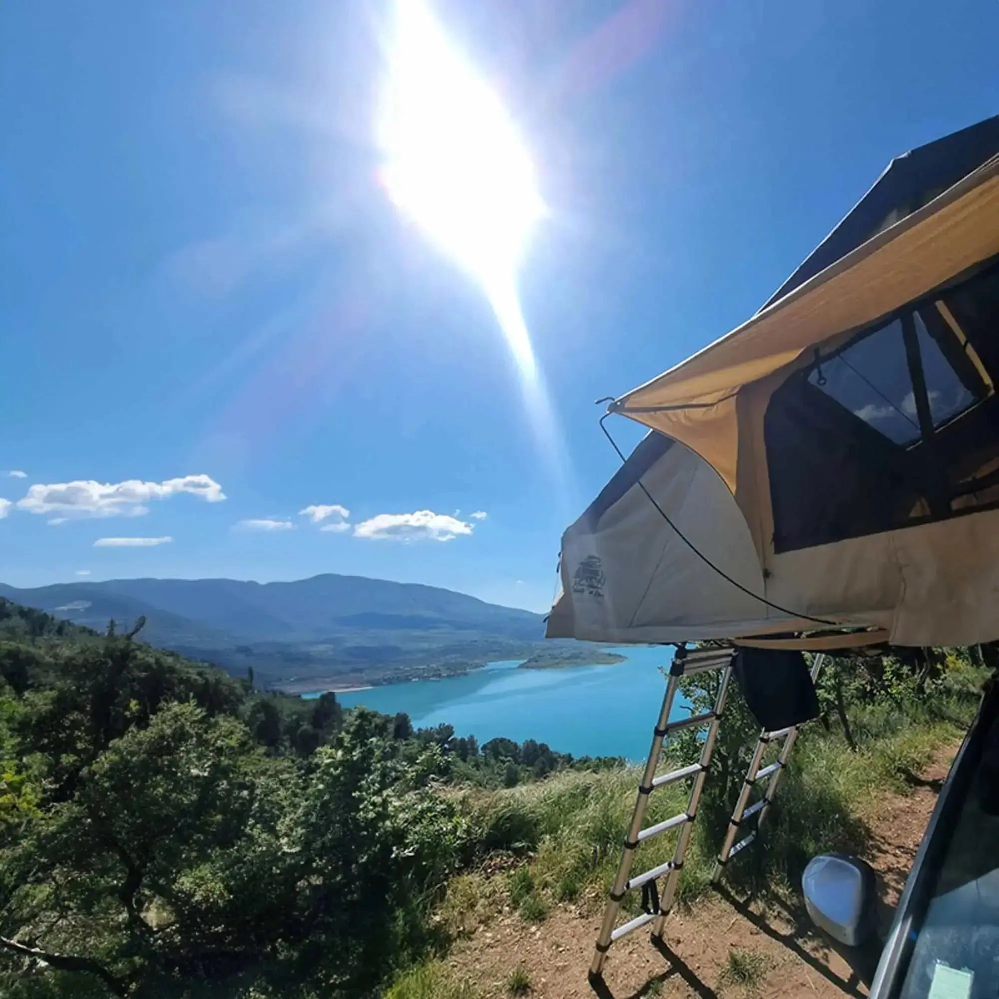 Roof tent setup with a scenic view of a lake and mountains in bright sunlight.