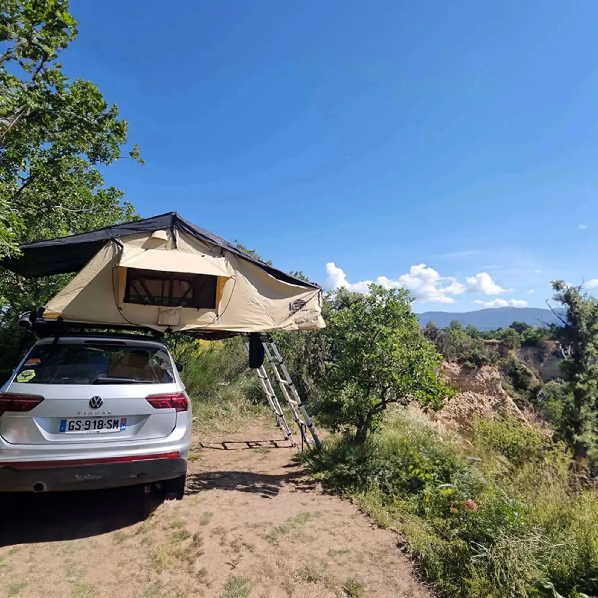 Roof tent set up on a car, beige with black top, parked in a scenic outdoor location with trees and blue sky.