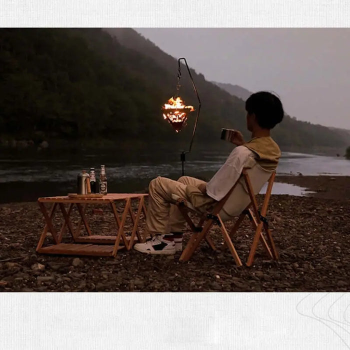 Scène de camping avec une personne assise près d'une rivière, avec une suspension de feu de camp triangulaire et une table en bois pour griller.