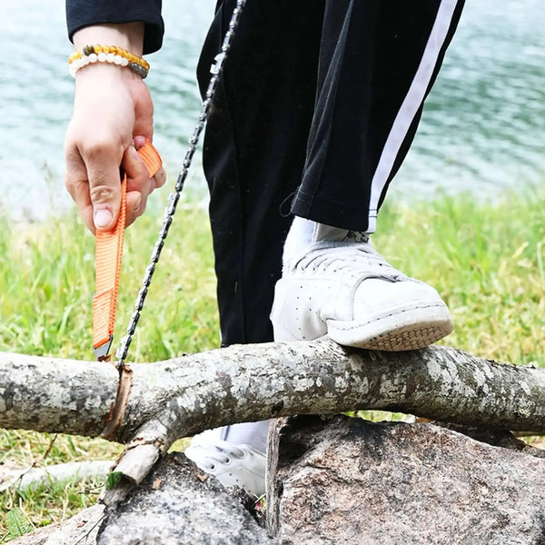 Une personne utilisant une scie à main portable pour camping avec des poignées orange pour couper une bûche au bord d'un lac. Idéale pour une coupe du bois rapide et sans ampoules.