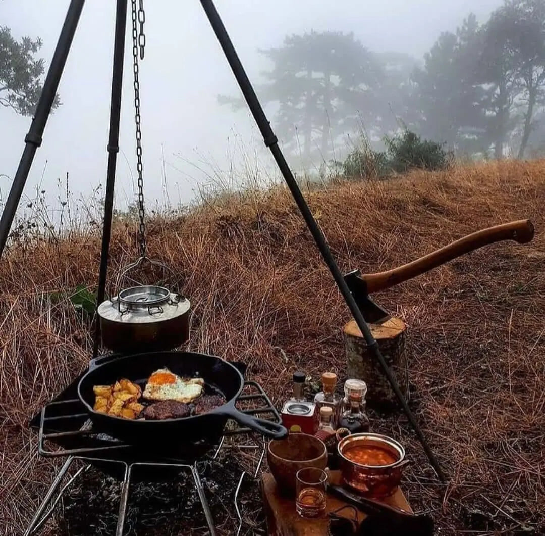 Poêle en fonte de 26cm au camping, cuisinant sur feu de camp, paysage brumeux, ustensiles de cuisine en plein air.