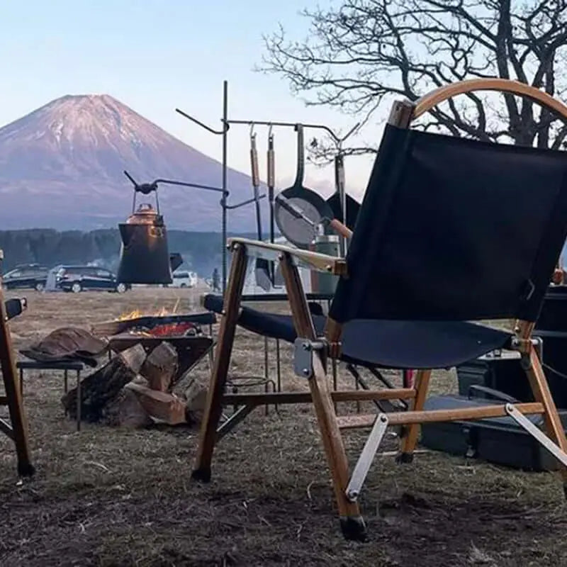 Chaise de camping pliable en bois noire près d'un feu de camp, avec vue sur une montagne au loin, parfaite pour activités de plein air.