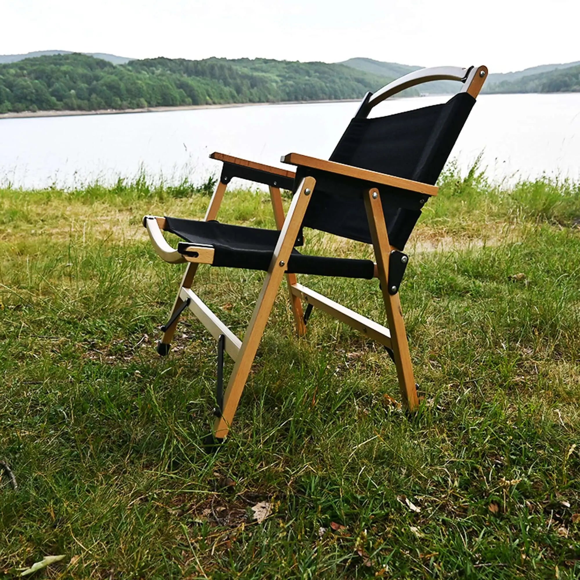Black foldable wooden camping chair on grassy lakeside, perfect for outdoor relaxation, featuring durable 600D Oxford fabric seat.