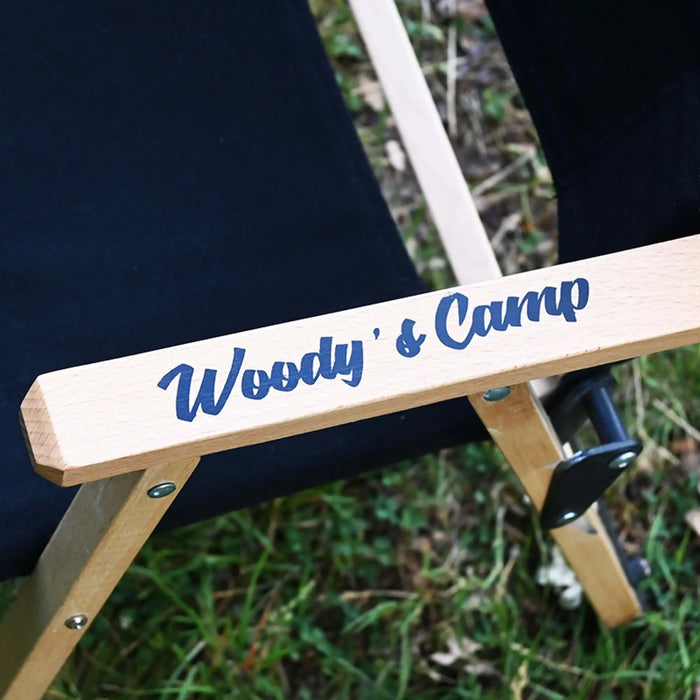 Gros plan sur une chaise de camping en bois noir pliable avec le texte "Woody's Camp", posée sur une pelouse à l'extérieur.