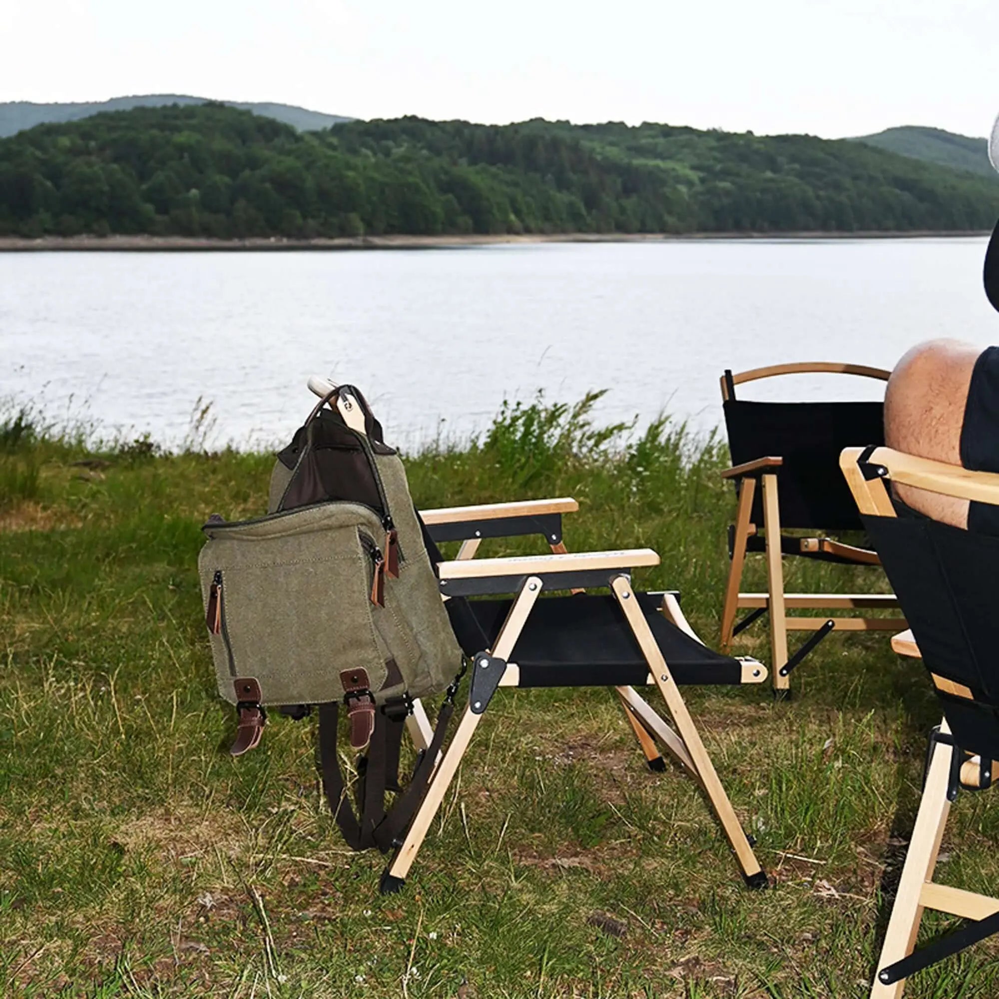 Chaise de camping pliante en bois noir avec sac à dos sur le bord d'un lac verdoyant