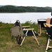 Chaise de camping pliante en bois noir avec un sac à dos au bord d'un lac verdoyant.