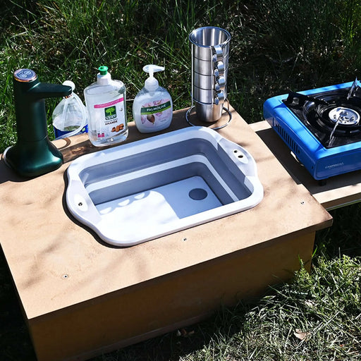 Évier pliable en silicone sur une table de camping avec des ustensiles et un réchaud à gaz, idéal pour la cuisine en plein air.