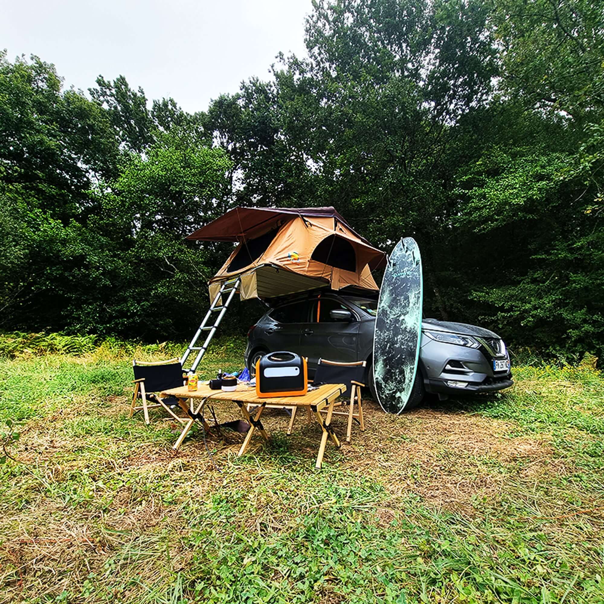Camping setup with rooftop tent on a car, folding table, chairs, and a surfboard in a forest setting.