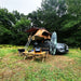 Installation de camping avec une tente de toit sur une voiture, une table pliante, des chaises et une planche de surf dans un cadre forestier.