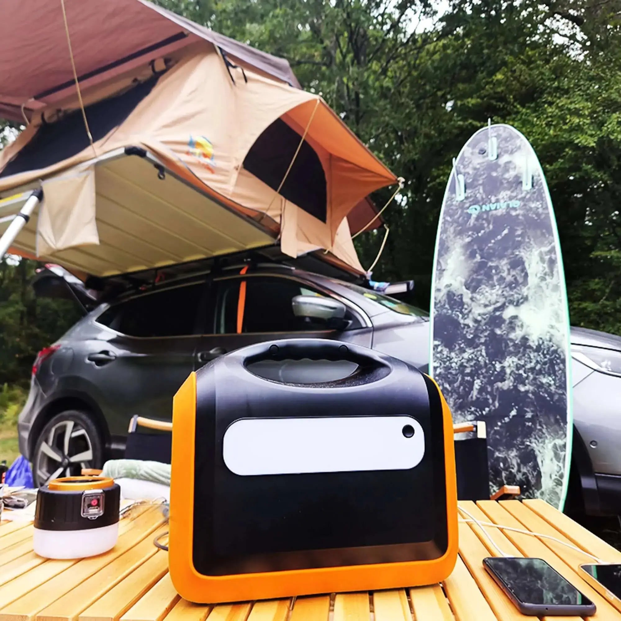Portable 1200W battery kit with solar panels, camping gear, and a surfboard next to an SUV with a rooftop tent in a forest setting.