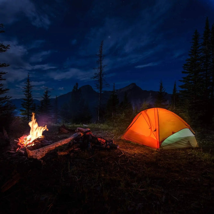 Tente de camping illuminée et feu de camp sous un ciel étoilé dans un cadre forestier.