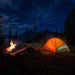 Tente de camping illuminée et feu de camp sous un ciel étoilé dans un cadre forestier.