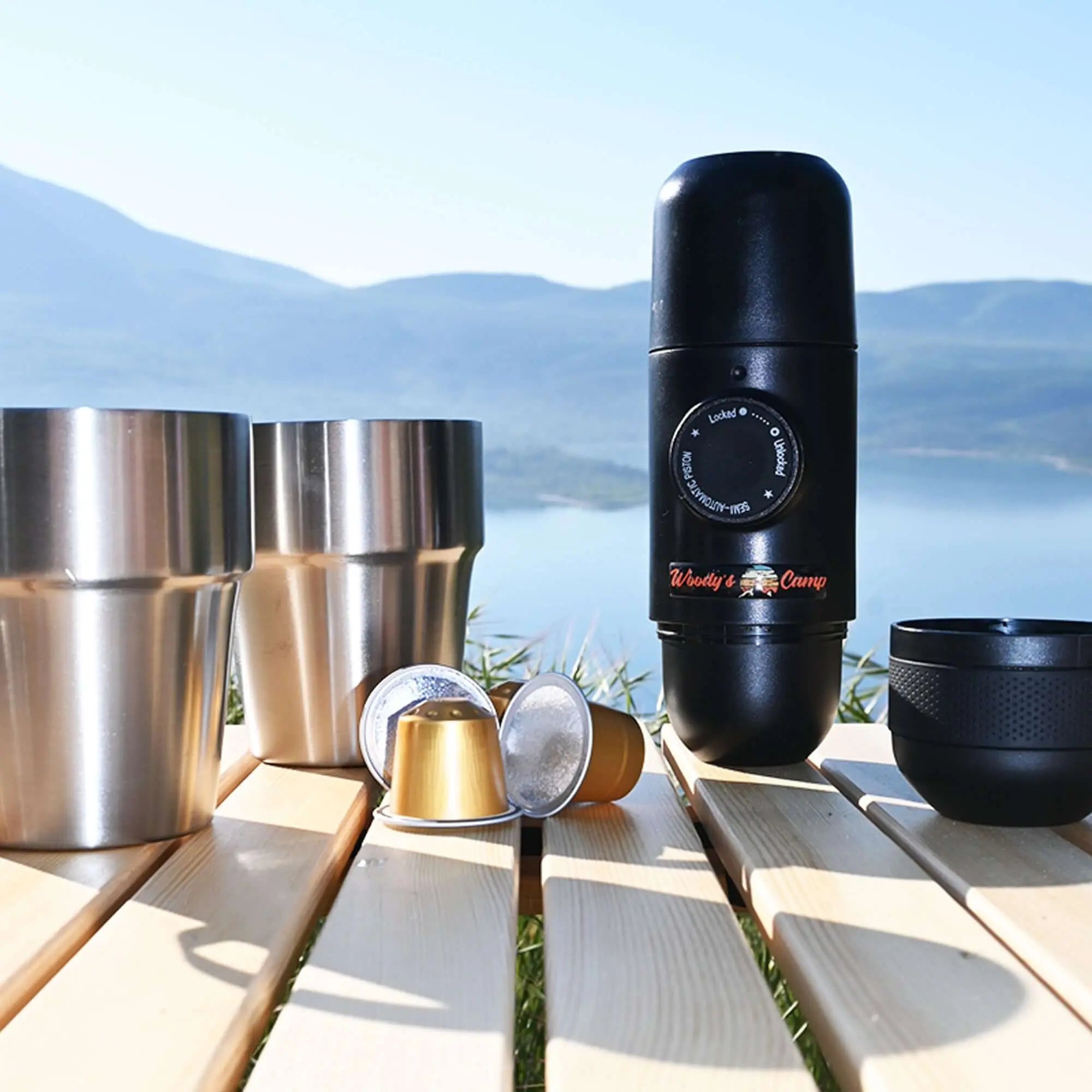 Portable Nespresso coffee maker with capsules on a picnic table near a scenic lake view, perfect for camping enthusiasts.