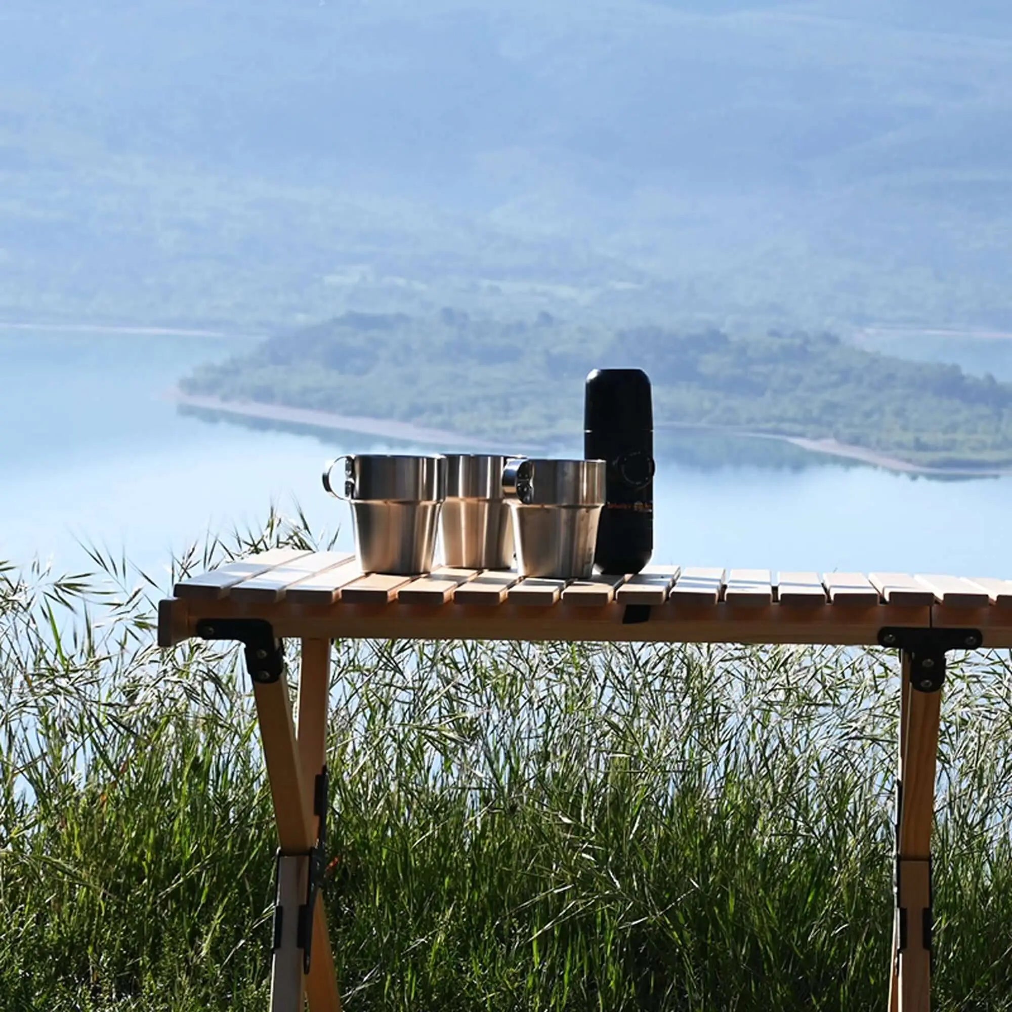 Portable espresso machine on a camping table with scenic lake view, ideal for outdoor coffee lovers.