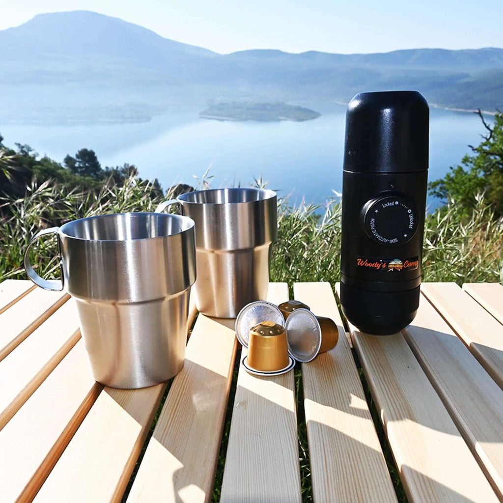 Portable espresso capsule machine with two stainless steel cups and capsules on a picnic table overlooking a scenic lake and mountains.
