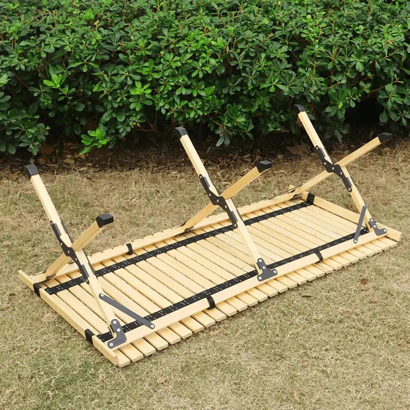 Foldable wooden camping table with sturdy hinges, partially unfolded, on a grassy area with a hedge in the background.
