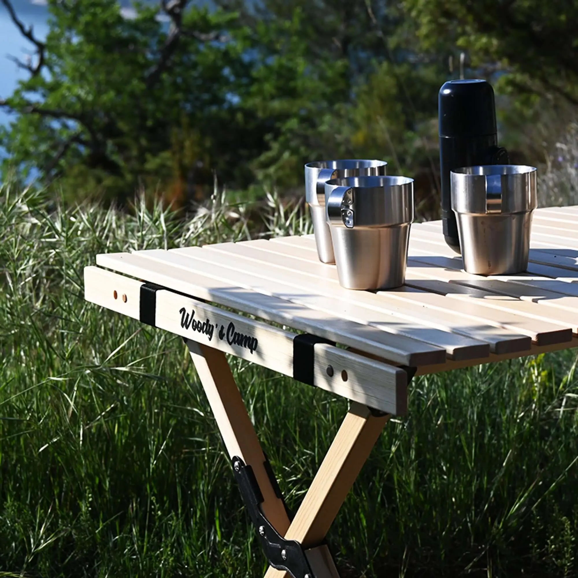 Table en bois pliable pour camping avec tasses en acier inoxydable, idéale pour repas en plein air, vue sur fond de nature.