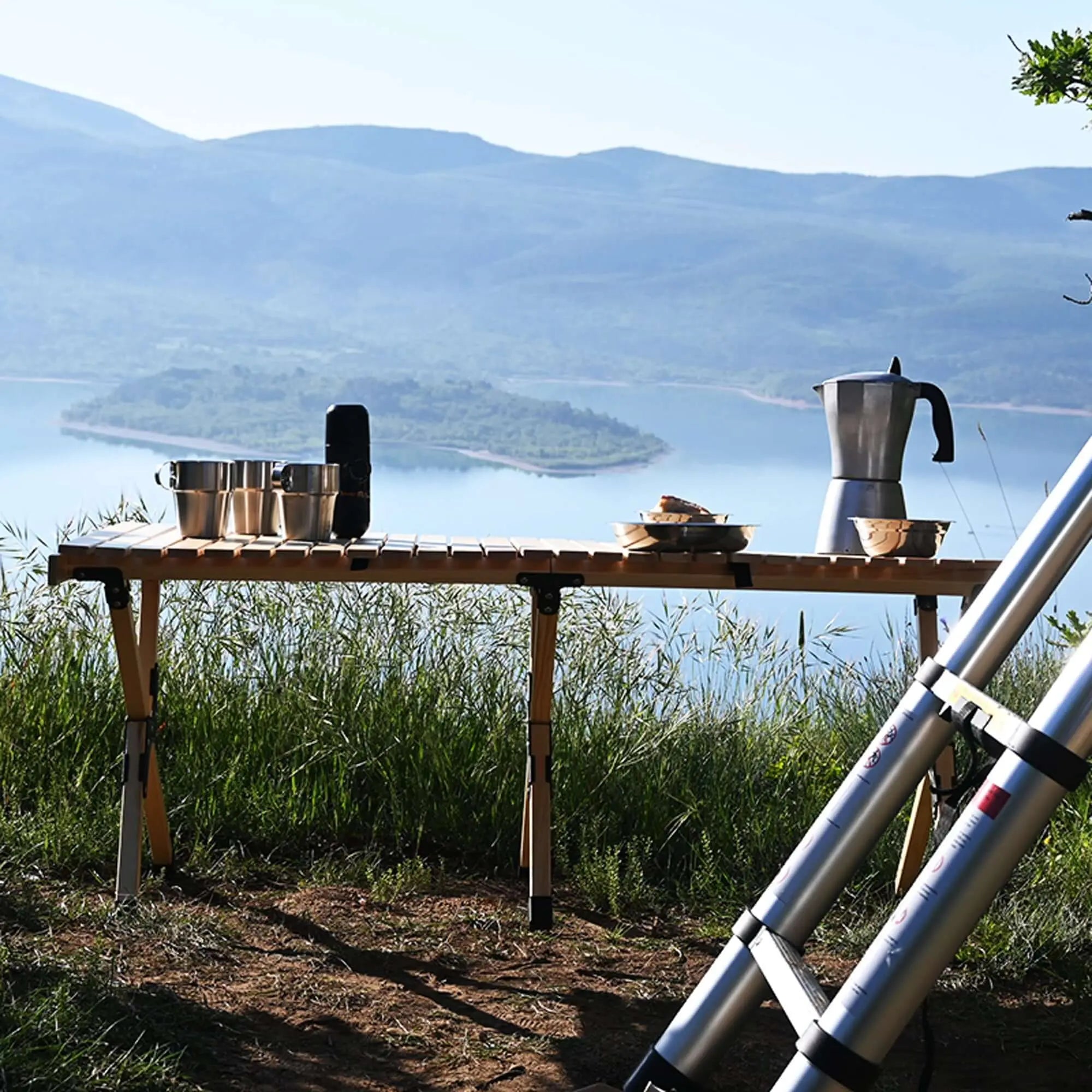 Table en bois pliable pour camping avec vaisselle sur fond de lac et montagnes, idéale pour repas en plein air, robuste et compacte.
