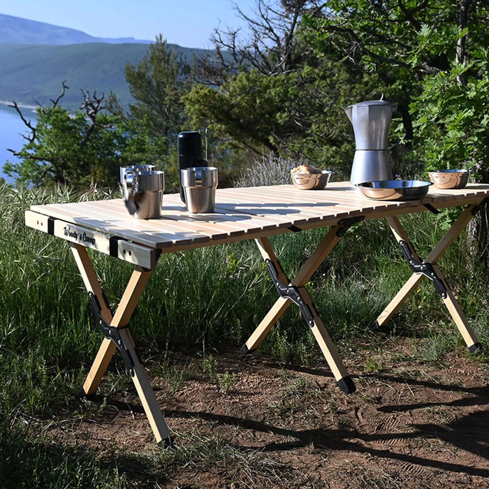 Table de camping en bois pliable avec vaisselle et cafetière dans un cadre extérieur pittoresque, conçue pour la commodité et la durabilité.