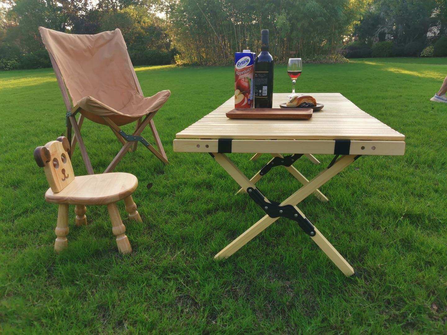 Table en bois pliable pour camping avec accessoires de pique-nique sur gazon, entourée de chaises pliantes, idéale pour repas en plein air.