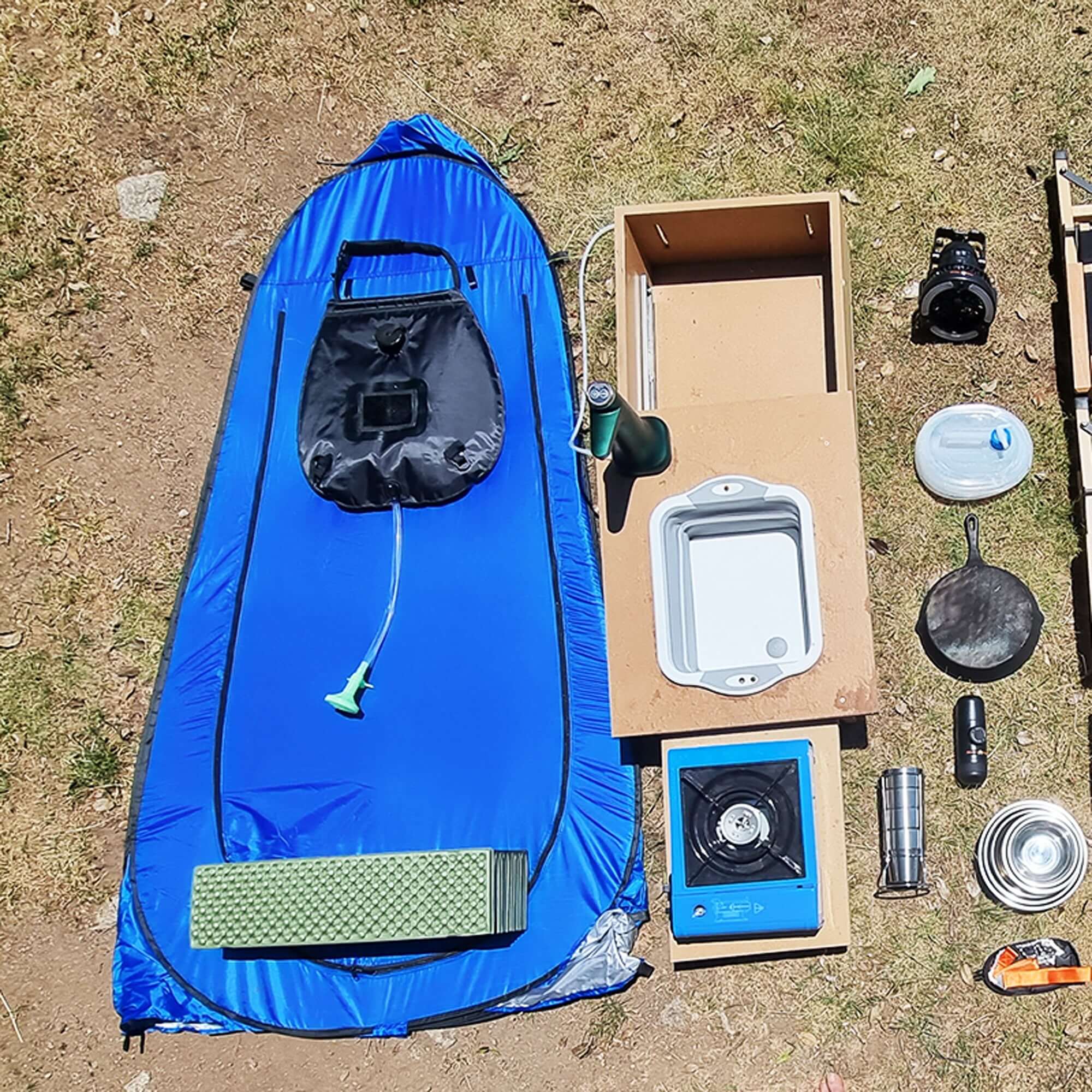 Pliable outdoor camping shower tent with camping gear on grass, including stove, pan, and water container, ideal for travel and adventure.